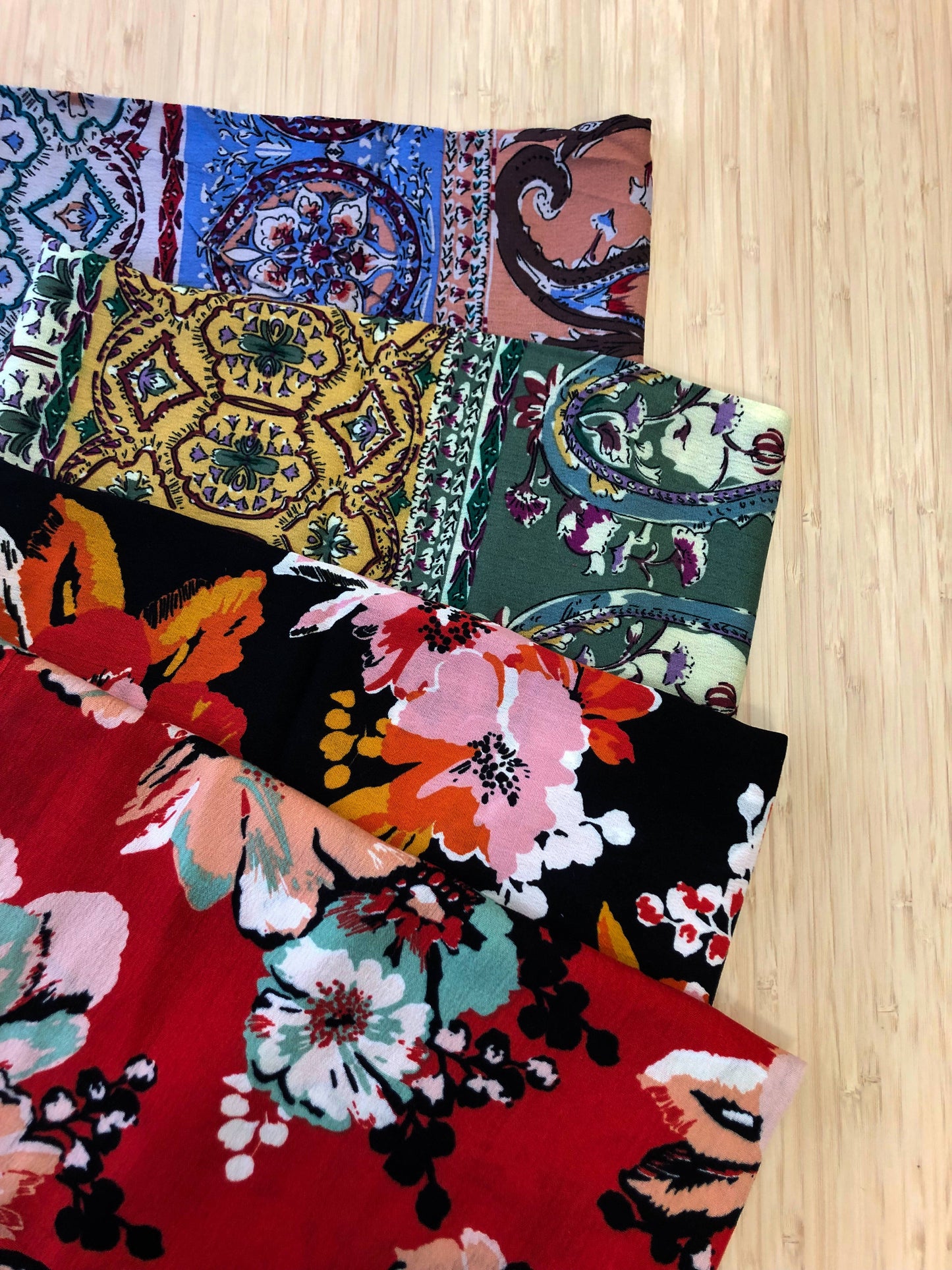 Photo of 4 different multicoloured floral prints of viscose fabric folded and stacked. photographed from above on a wooden table.