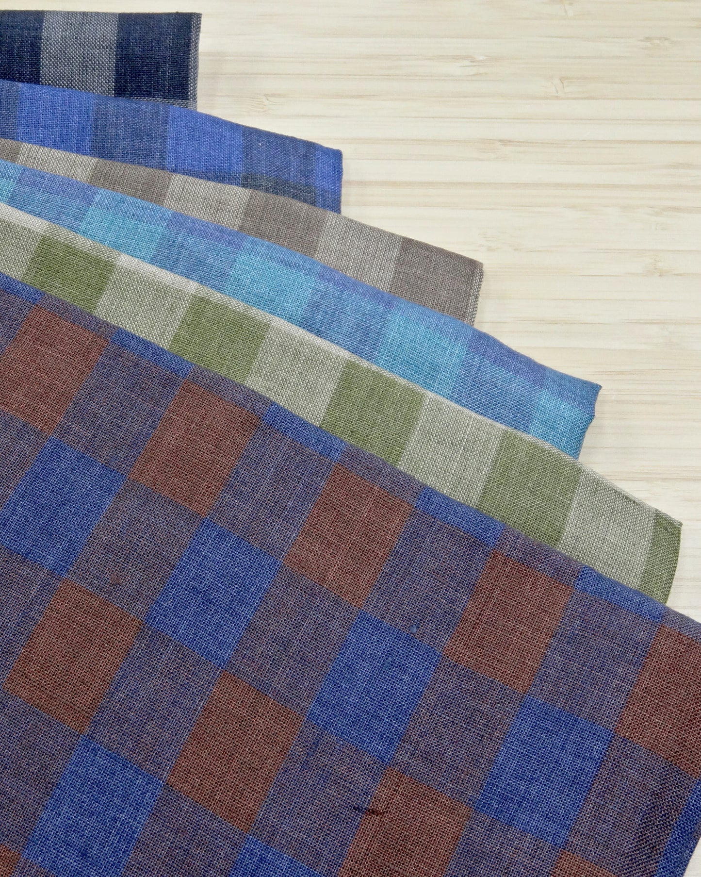 Six different colours of colourful yarn dyed check fabrics in a stack on a wooden table.
