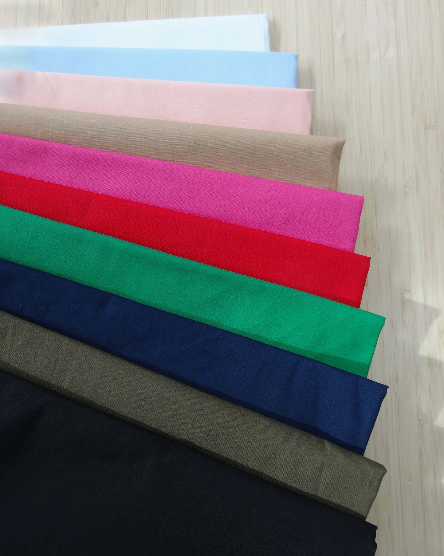 Photo of 10 different colours of 120gsm cotton poplin fabrics folded and stacked. photographed from above on a wooden table.