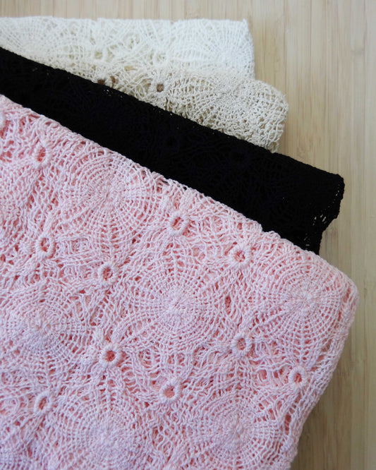 Photo of crochet style macrame lace fabrics folded and stacked in pink, black, beige, and white photographed from above on a wooden table