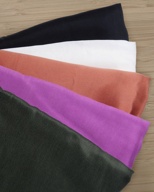 Photo of 5 different colours of Cupro Rayon blended satin fabric folded and stacked.  photographed from above on a wooden table.