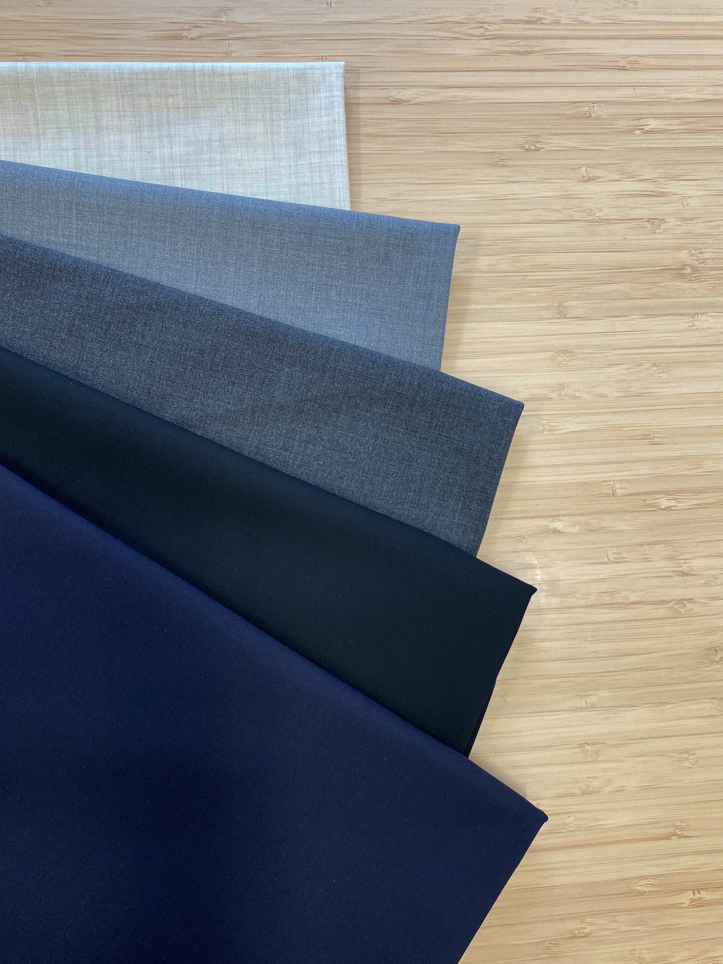 Photo of 5 different colours of Poly Rayon Suiting Fabric folded and stacked. photographed from above on a wooden table.
