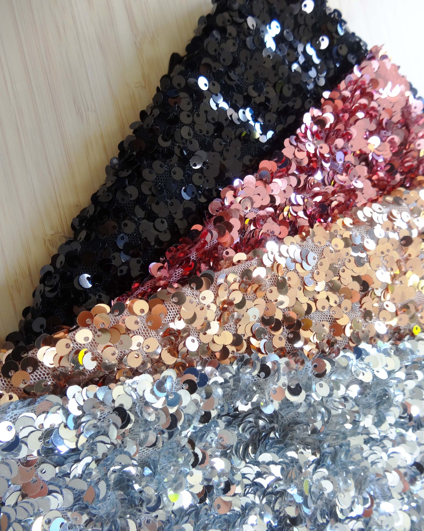 Photo of 4 sequin fabrics folded and stacked in silver, gold, pink, and black photographed from above on a wooden table.