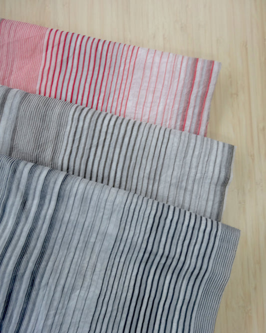 Photo of 3 different colours of silk cotton striped fabrics folded and stacked in black stripe on white base, brown stripe on white base, and red stripes on white fabric.  photographed from above on a wooden table.
