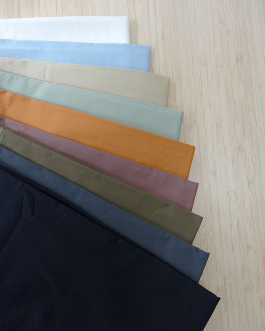 Photo of 9 different colours of 150gsm cotton stretch poplin fabrics folded and stacked.  photographed from above on a wooden table.