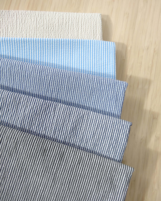 Photo of 5 different colours of 145gsm striped seersucker cotton polyester spandex fabrics folded and stacked.  photographed from above on a wooden table.