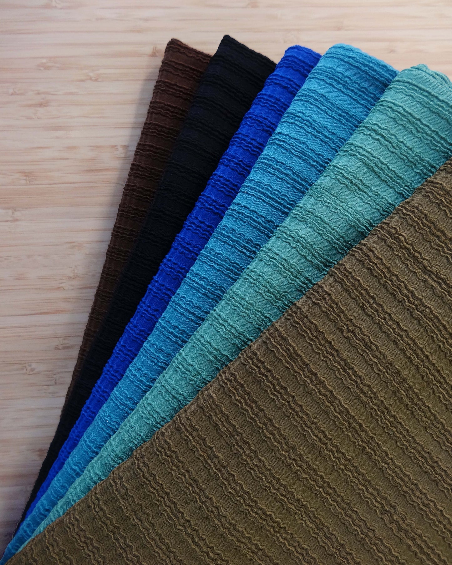 Photo of ribbed knit fabrics folded and stacked in khaki green, teal, mid blue, cobalt blue,, black, and chocolate brown photographed from above on a wooden table.