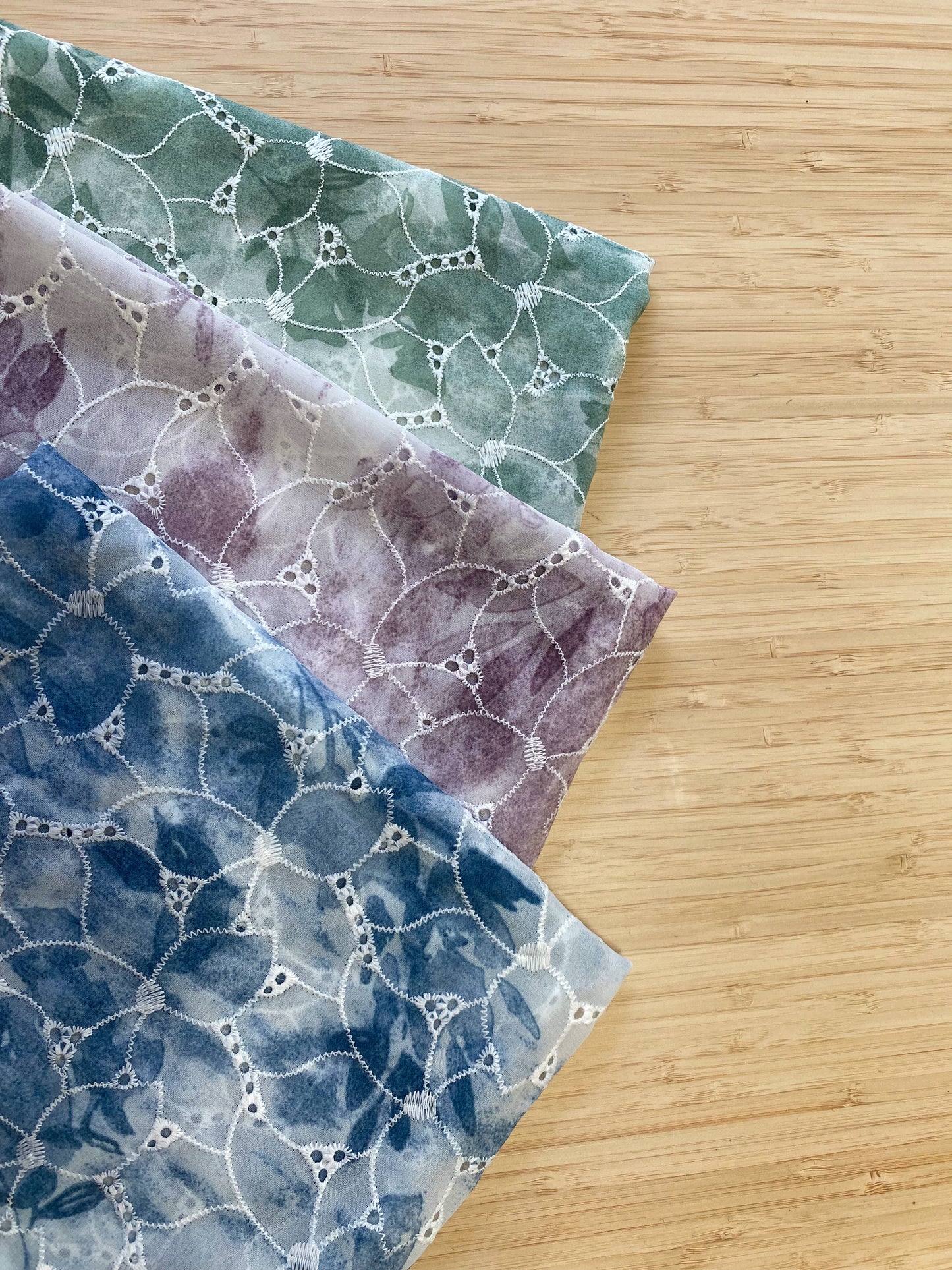 Photo of 3 different colours of Tie Dye Floral Embroidered fabric folded and stacked. photographed from above on a wooden table.