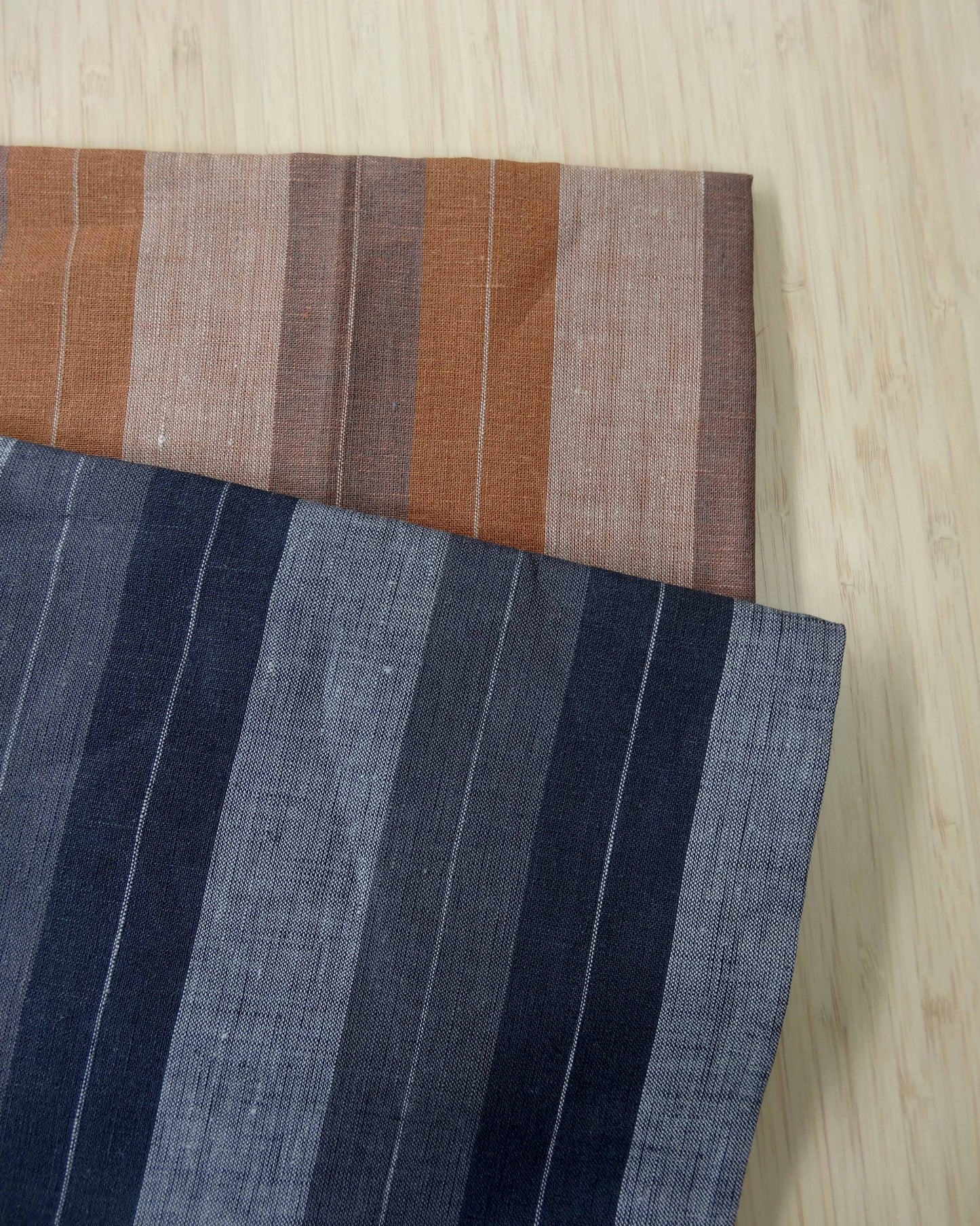 Photo of 2 different colours of striped linen fabrics folded and stacked. One is in orange tones and the other in blue tones. photographed from above on a wooden table.