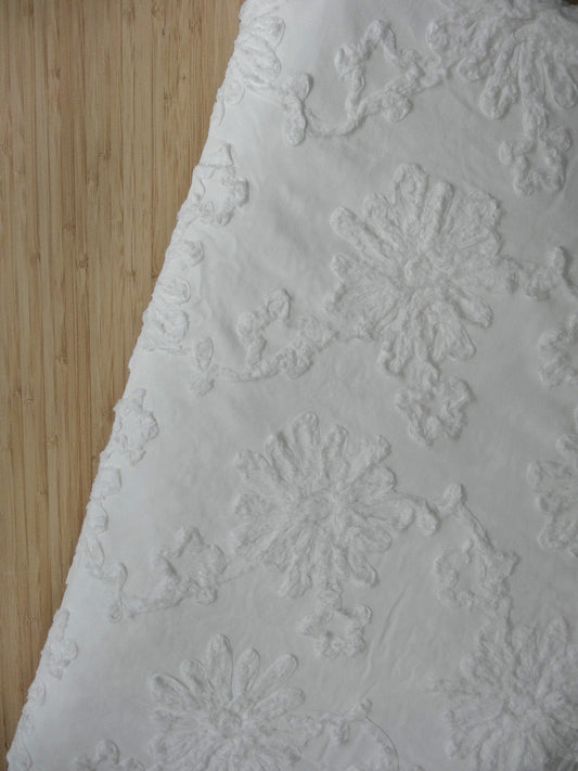 White 100% cotton fabric with white floral applique. Fabric is folded and photographed from above on a wooden table.