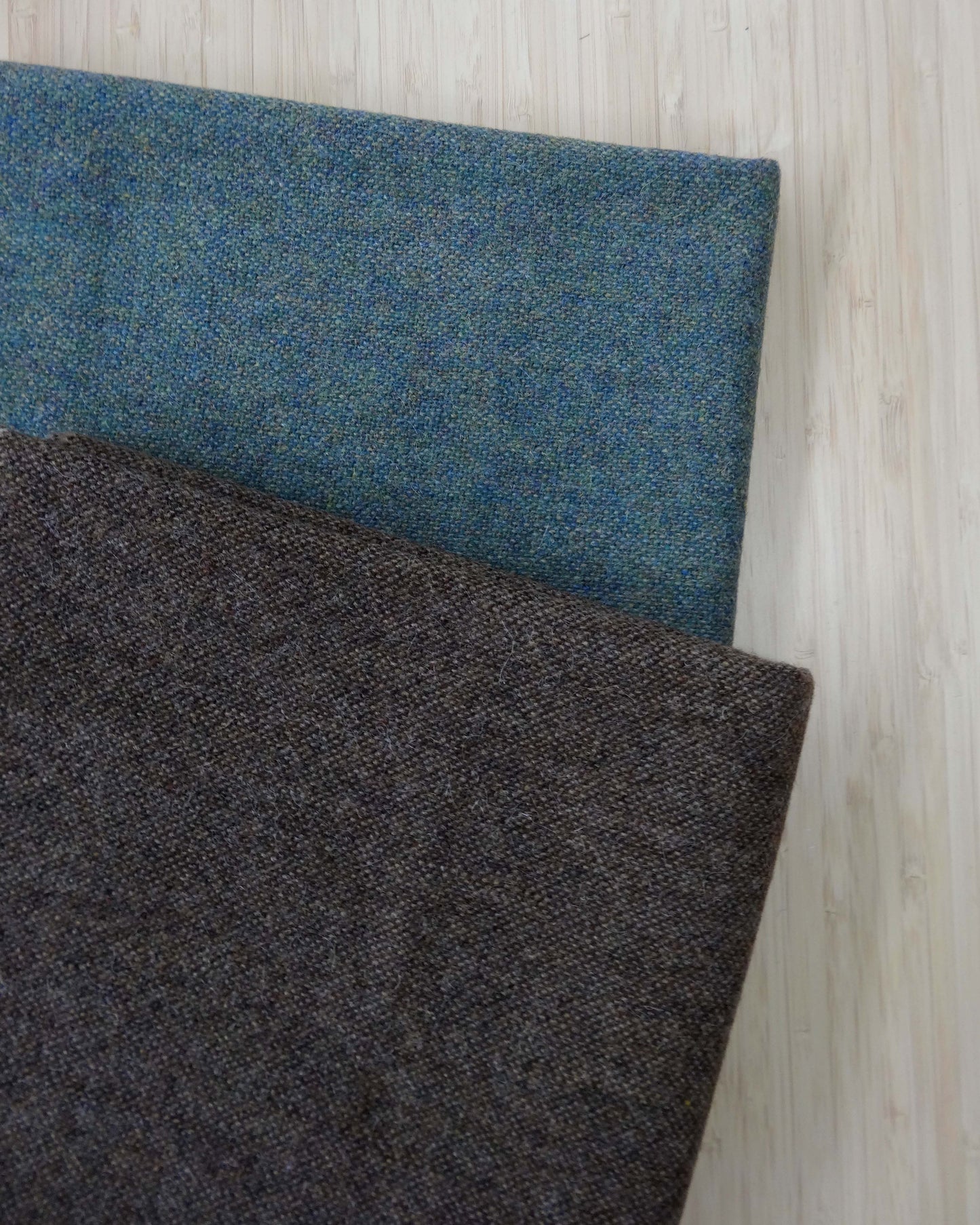 Two pieces of wool fabric folded and stacked. One is a mottled brow and the other is a mottled blue/teal tone. Photographed from above on a wooden table.