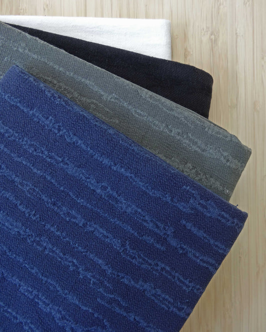 Photo of textured jacquard fabric short ends stacked in blue, khaki green, black, and white on a wooden table. photographed from above.