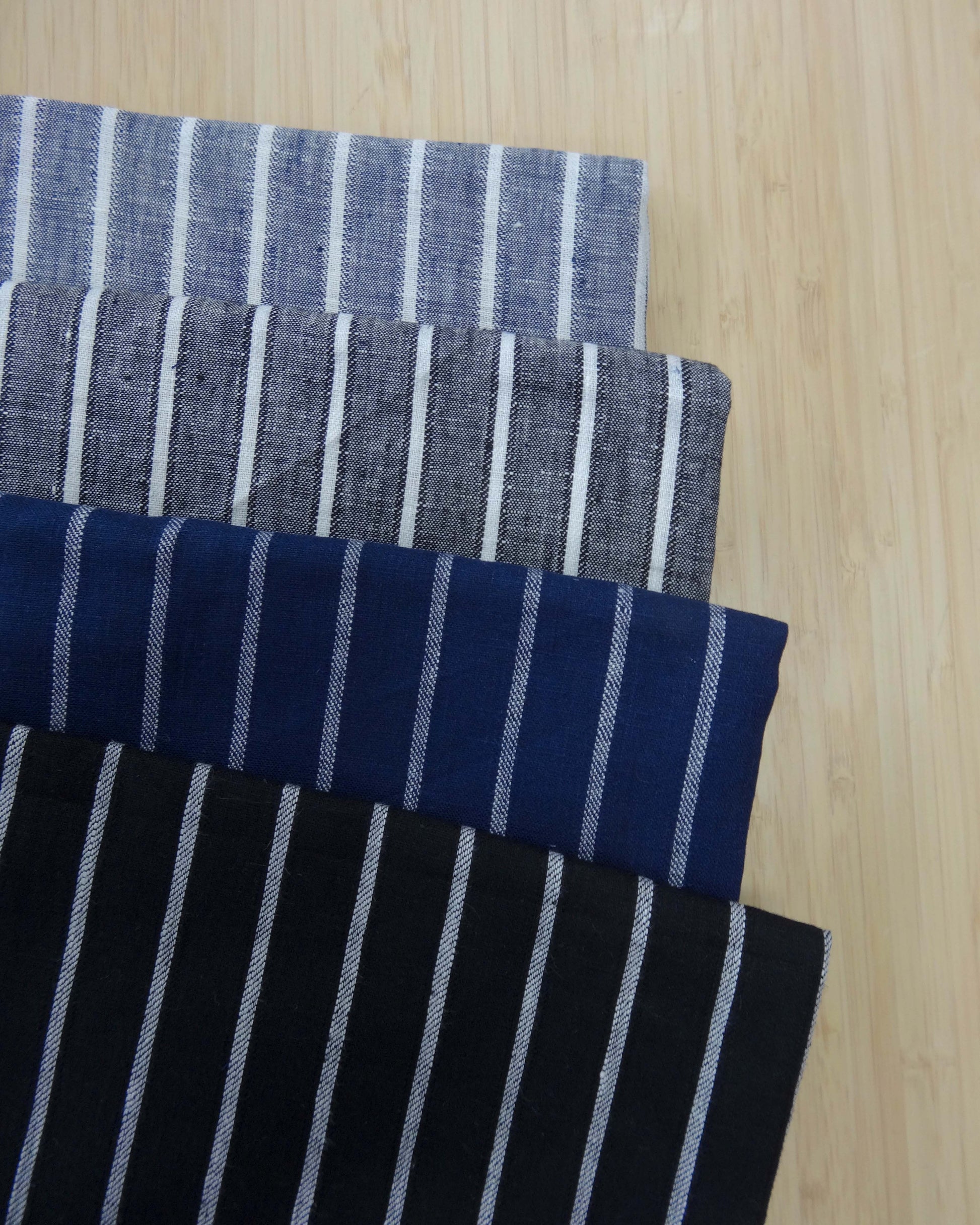 Photo of 4 different colours of 163gsm striped 100% linen fabrics folded and stacked.  photographed from above on a wooden table.