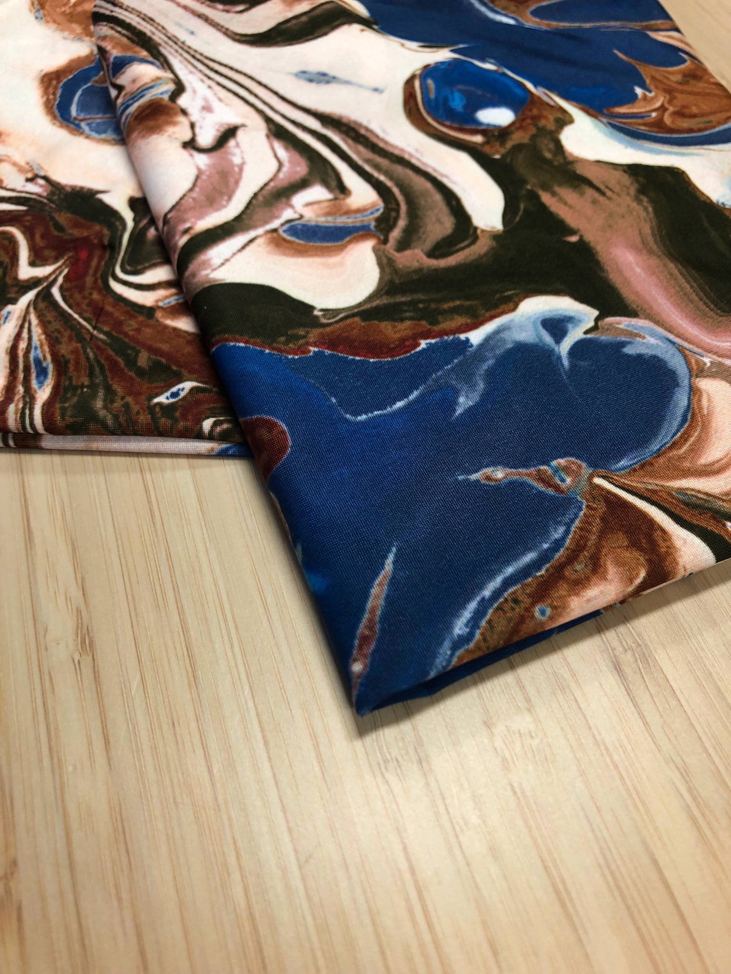 Photo of a printed jersey fabric, the print is an abstract swirling marble like design in blues, browns and whites, the fabric is folded and photographed on a wooden table
