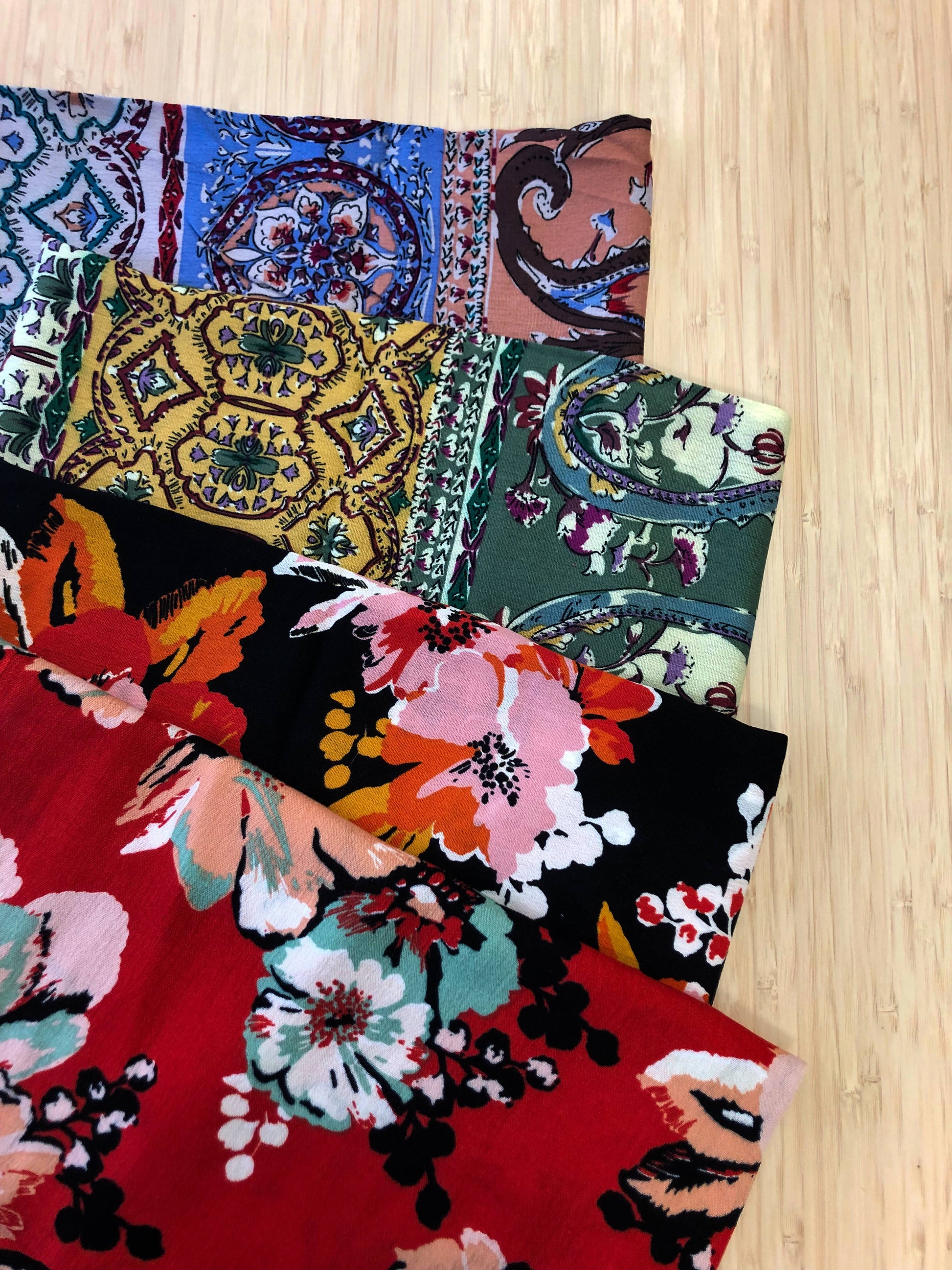 Photo of 4 different multicoloured floral prints of  viscose fabric folded and stacked. photographed from above on a wooden table.