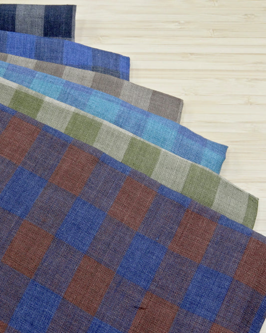 Six different colours of colourful yarn dyed check fabrics in a stack on a wooden table.