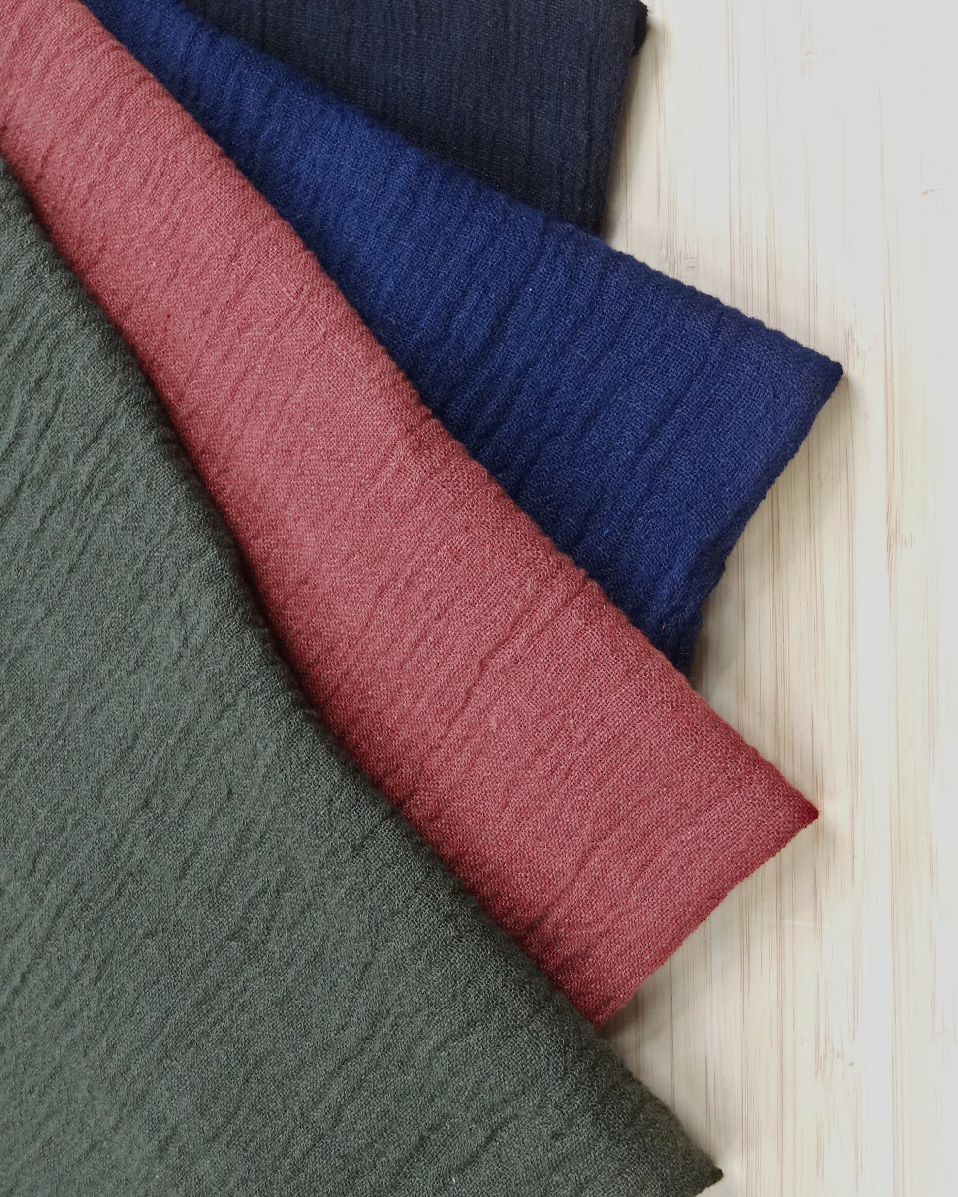 Four colours of fabric stacked and laid on a wooden table. The fabric is Khaki, rose, cobalt, and charcoal