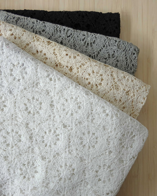 Photo of crochet style macrame lace fabrics folded and stacked in white, beige, grey and black photographed from above on a wooden table