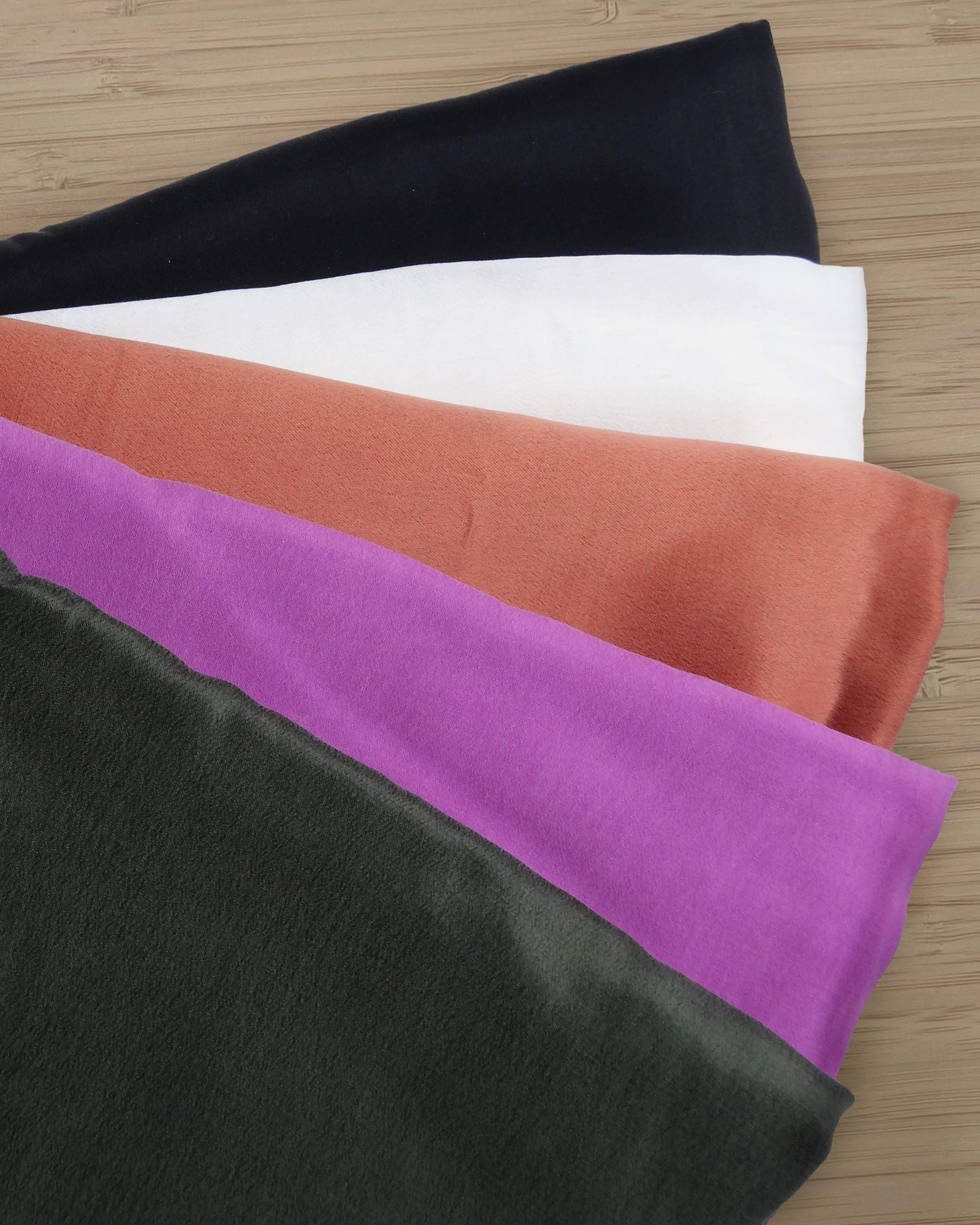 Photo of 5 different colours of Cupro Rayon blended satin fabric folded and stacked.  photographed from above on a wooden table.