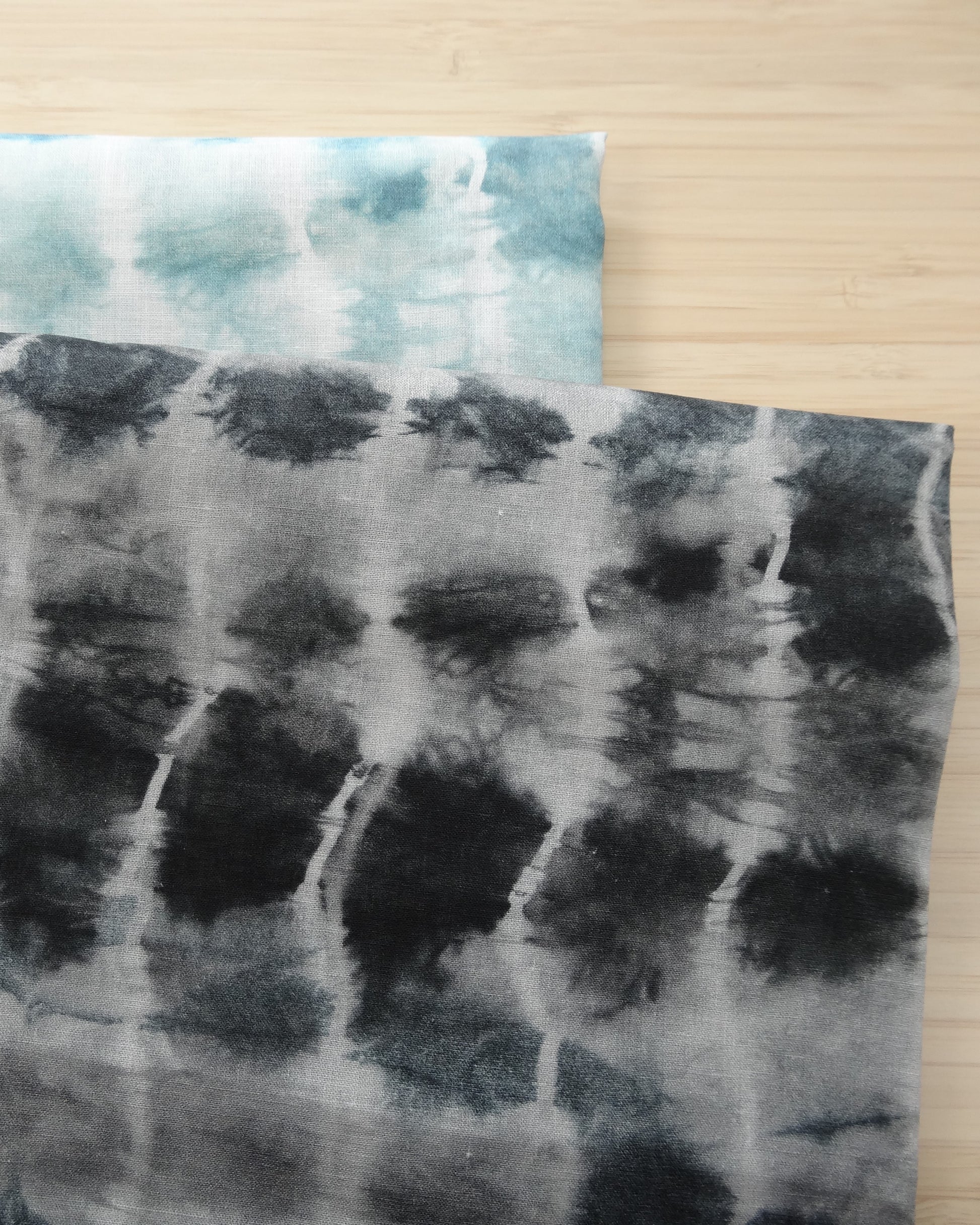 Photo of ripple design printed linen cotton blended fabric remnants stacked in light blue and black/greys  on a wooden table. photographed from above.
