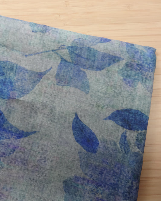 Photo of leafy floral digital print on lightweight ramie fabric remnants stacked in blue on a wooden table. photographed from above.