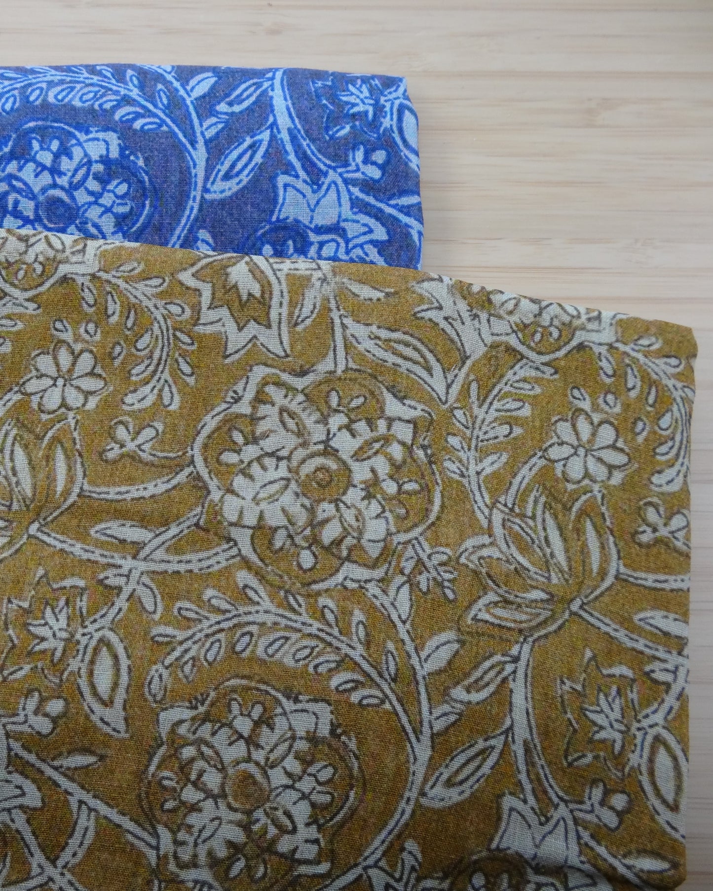 Photo of drawn floral digital print on lightweight ramie fabric remnants stacked in blue and mustard on a wooden table. photographed from above.