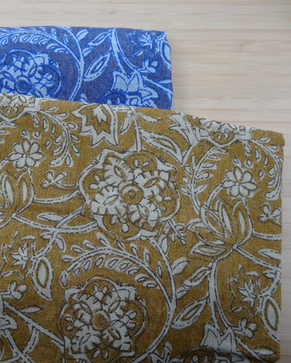 Photo of drawn floral digital print on lightweight ramie fabric remnants stacked in blue and mustard on a wooden table. photographed from above.