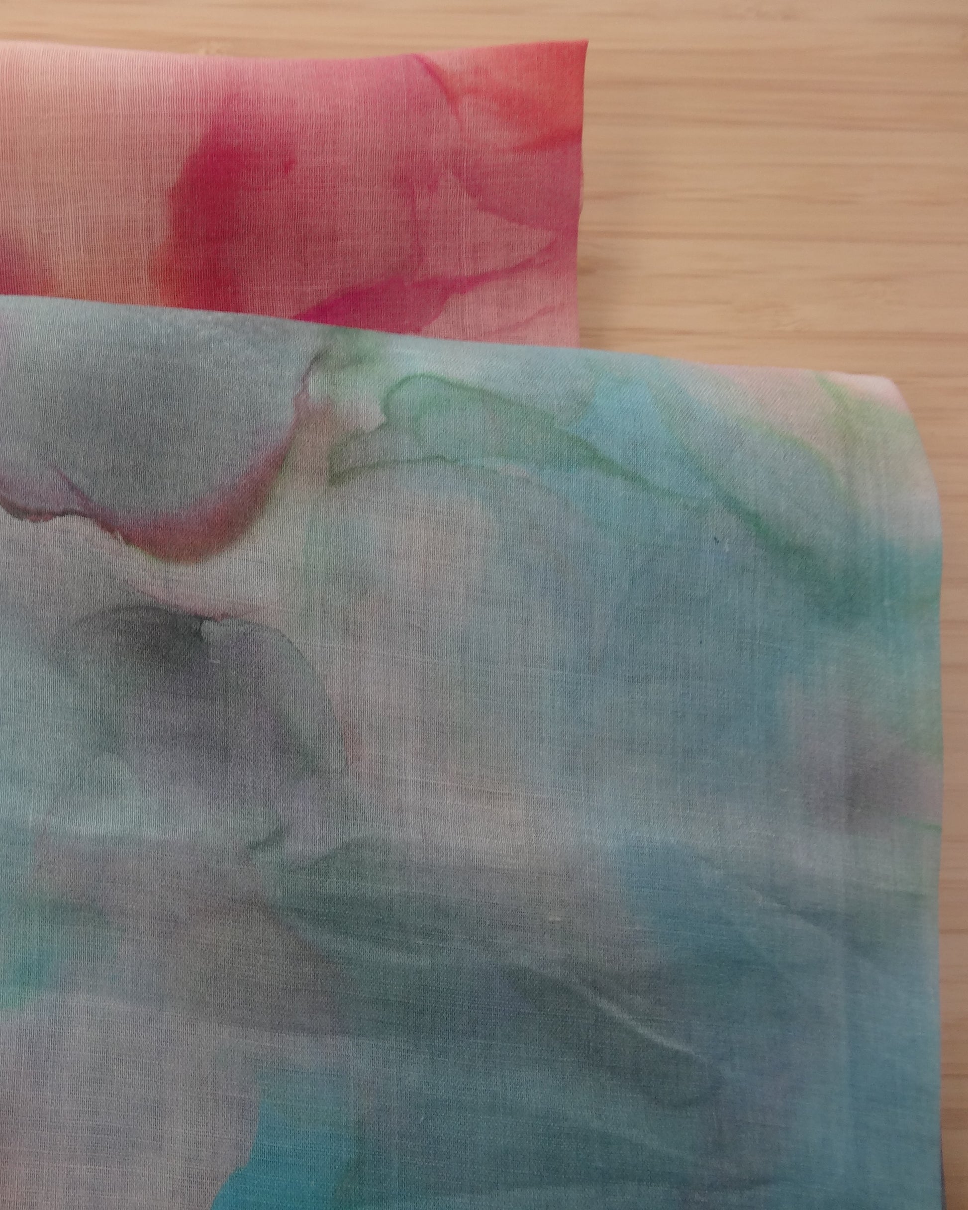 Photo of abstract watercolour abstract digital print on lightweight ramie fabric remnants stacked in blues and pinks on a wooden table. photographed from above.