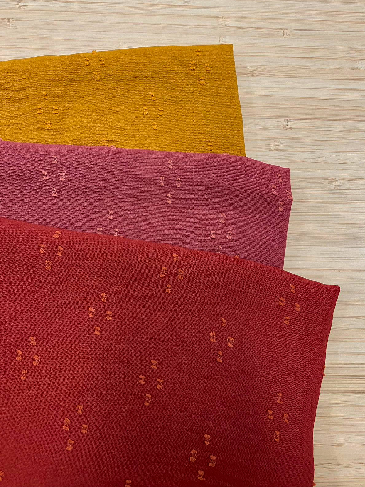 Photo of 3 different colours of Spot detail Jacquard fabric folded and stacked. photographed from above on a wooden table.
