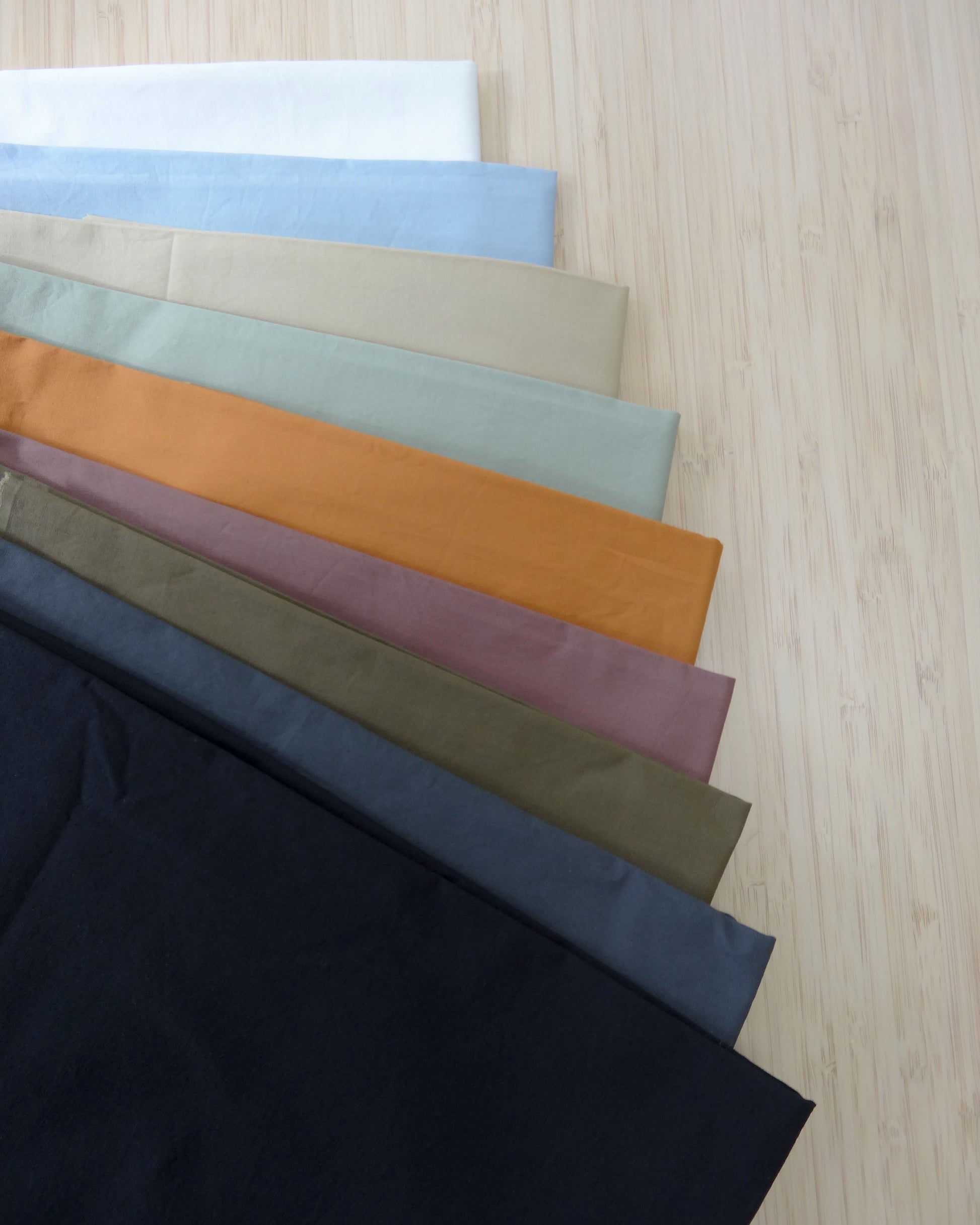Photo of 9 different colours of 150gsm cotton stretch poplin fabrics folded and stacked.  photographed from above on a wooden table.
