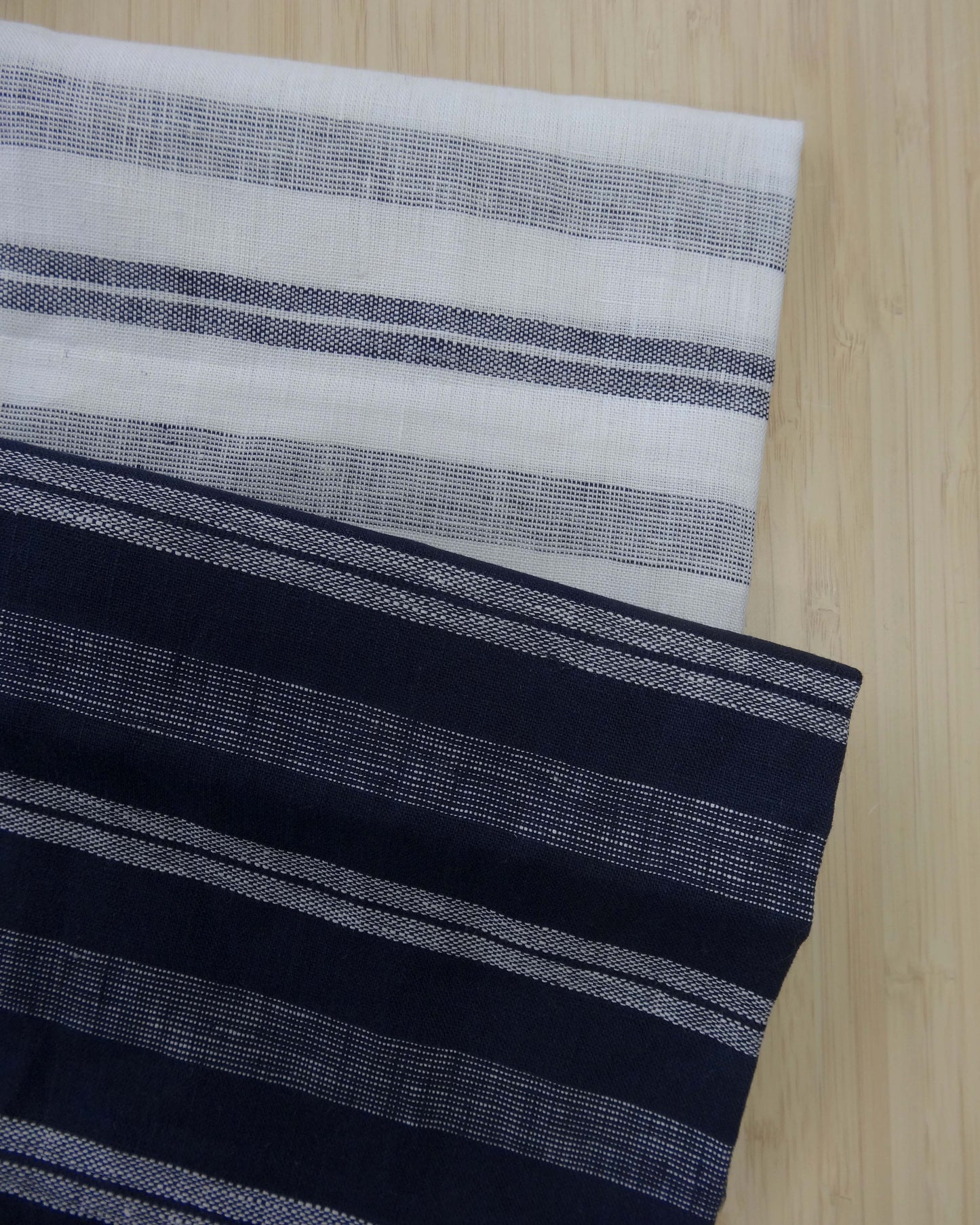 Photo of 2 different colours of 137gsm striped linen fabrics folded and stacked.  photographed from above on a wooden table.
