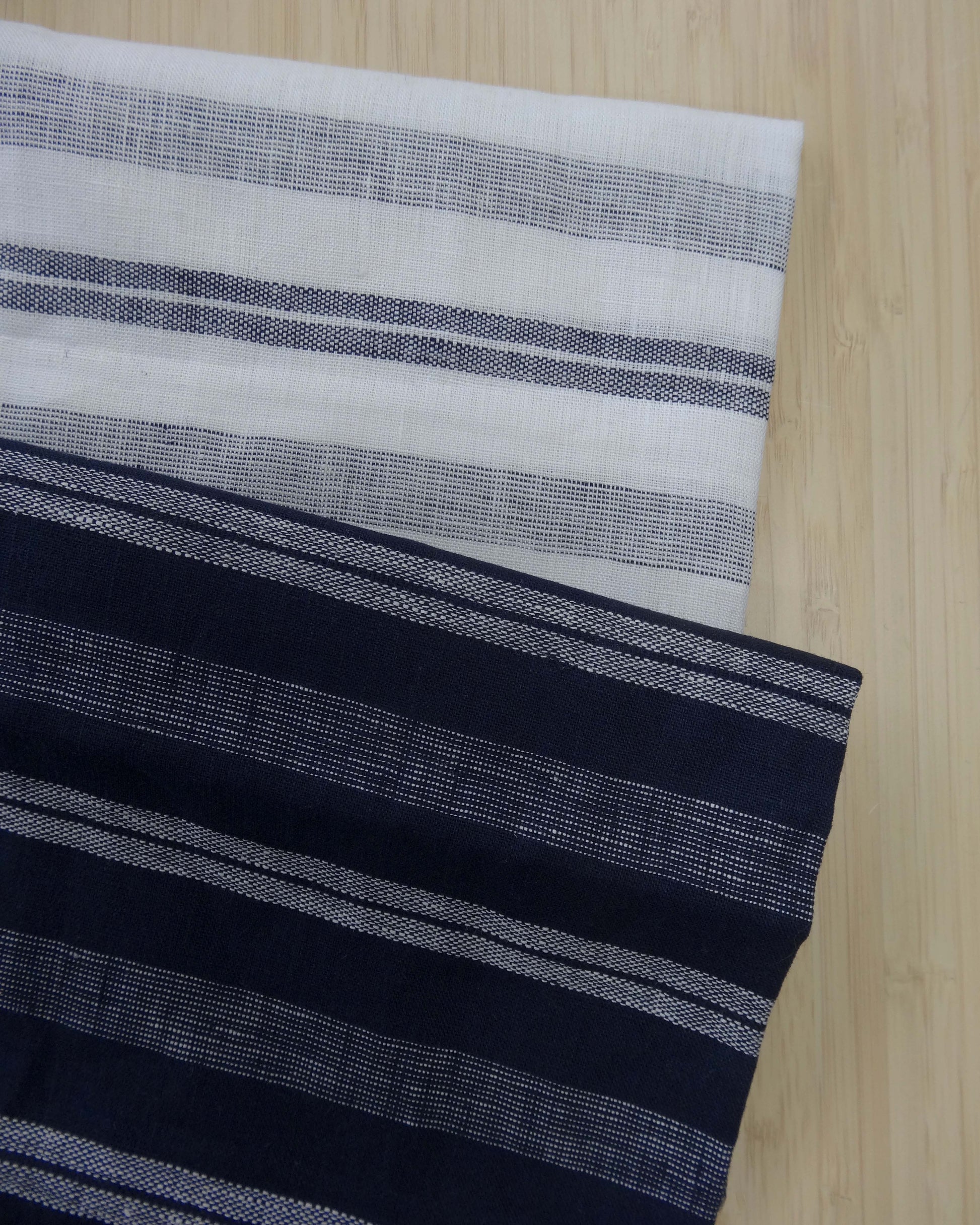 Photo of 2 different colours of 137gsm striped linen fabrics folded and stacked.  photographed from above on a wooden table.
