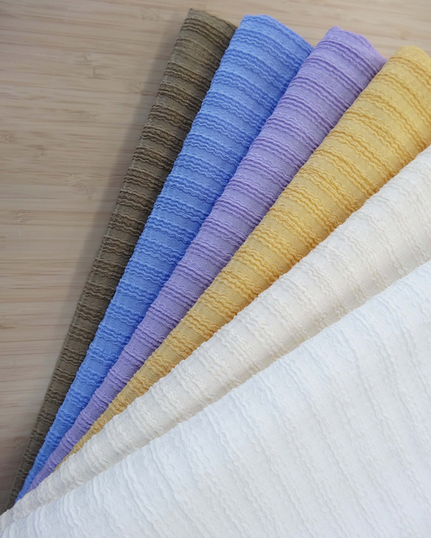 Photo of wavy ribbed knit fabrics folded and stacked in white, cream, yellow, purple, cornflower blue, and walnut brown photographed from above on a wooden table.