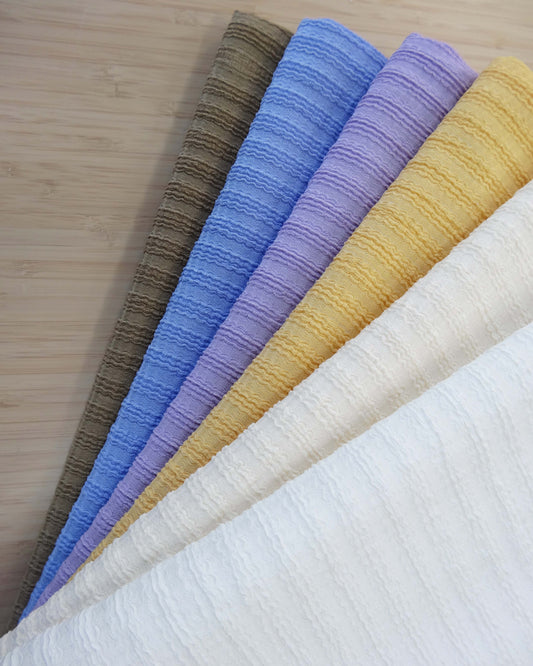 Photo of wavy ribbed knit fabrics folded and stacked in white, cream, yellow, purple, cornflower blue, and walnut brown photographed from above on a wooden table.