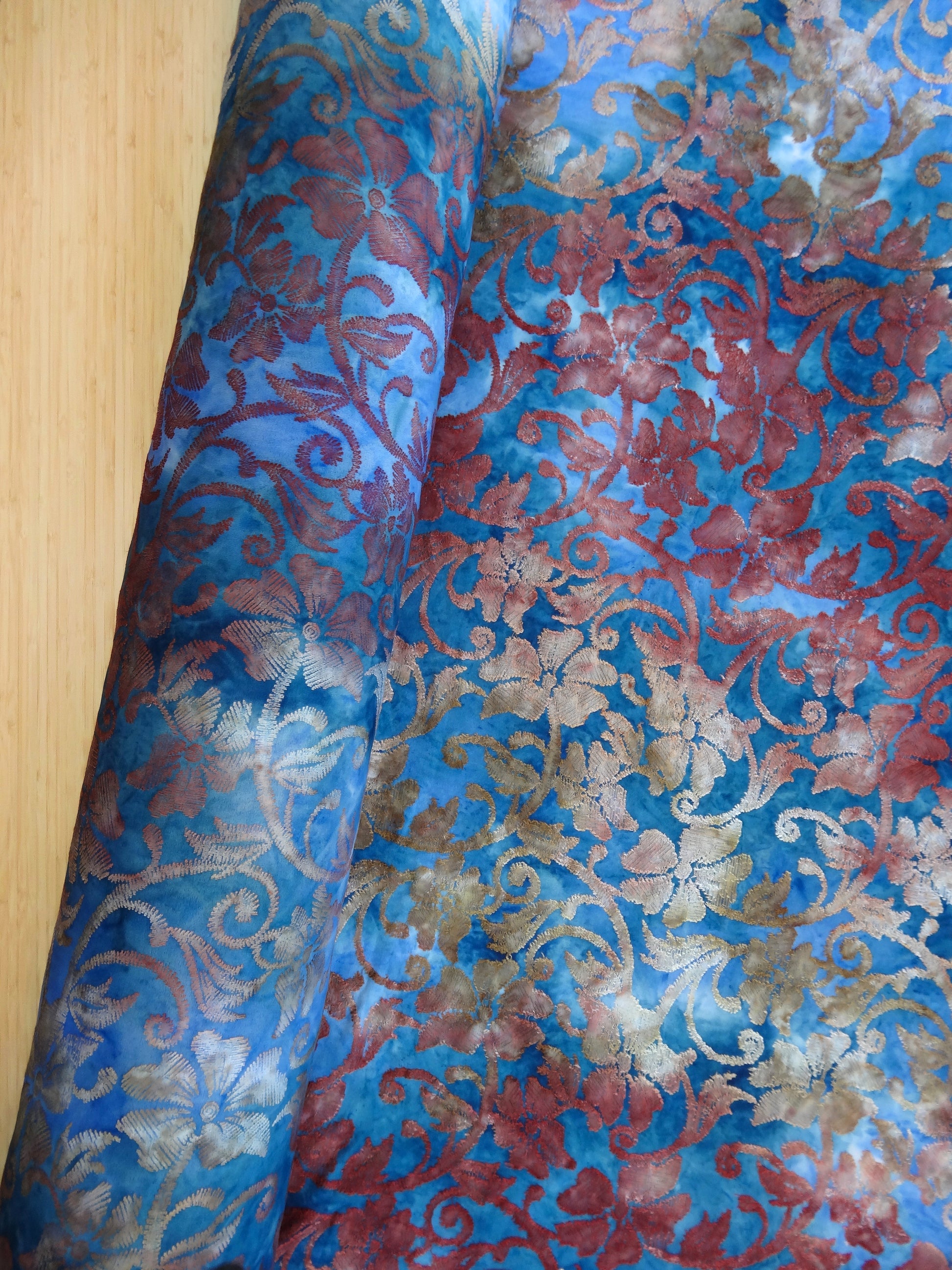 Blue based tie-dye fabric with red and ochre floral jacquard details. it is on a roll and partially unraveled from the roll. it is being photographed from above on a wooden table.