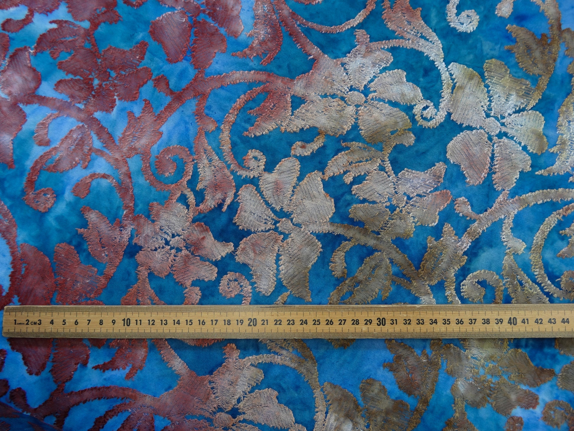 Blue based tie-dye fabric with red and ochre floral jacquard details. Close up photograph from above with wooden ruler used to show scale of the print