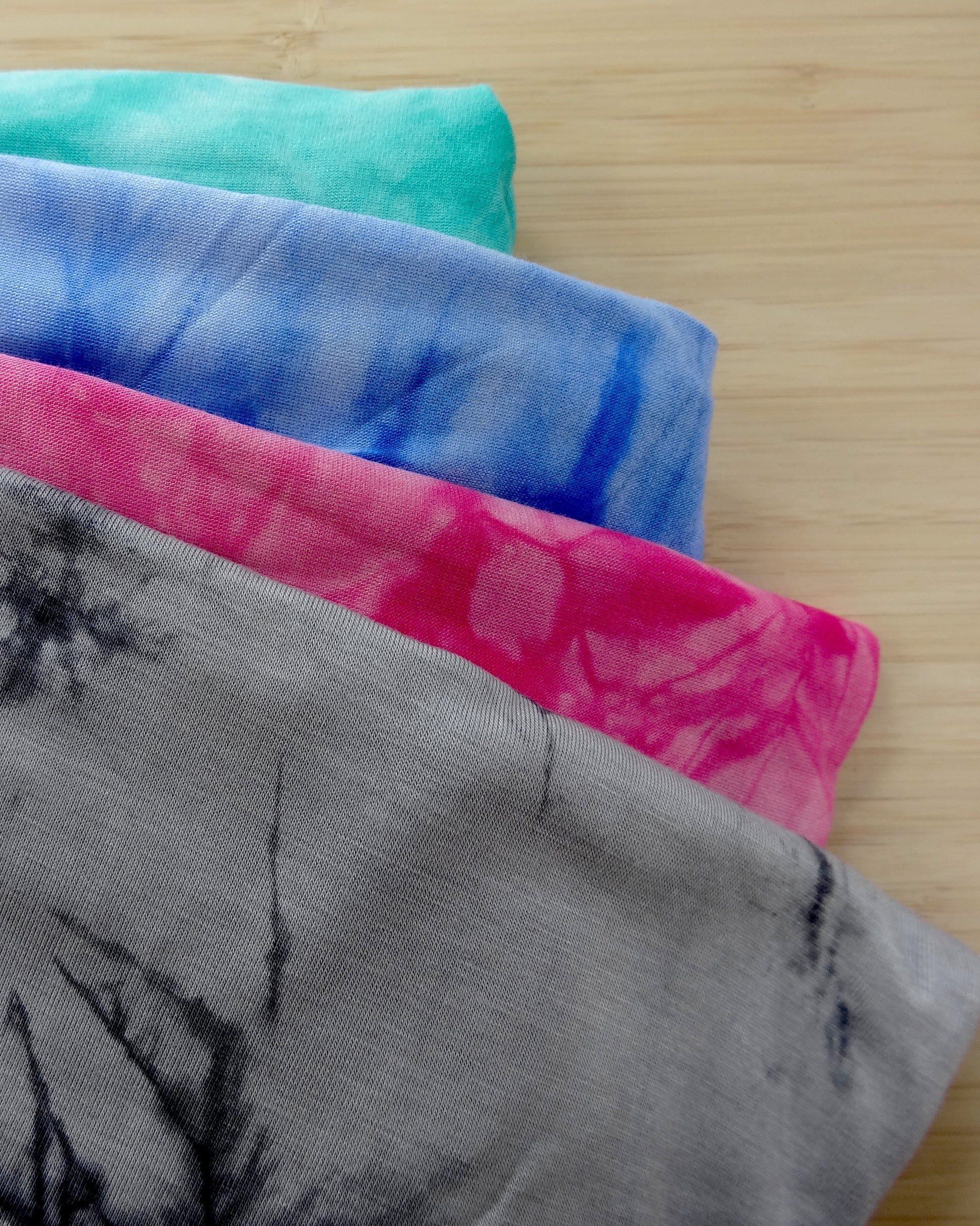 Four folded tie dyed rayon blended fabric swatches in teal, blue, pink, and gray on a wooden surface. 