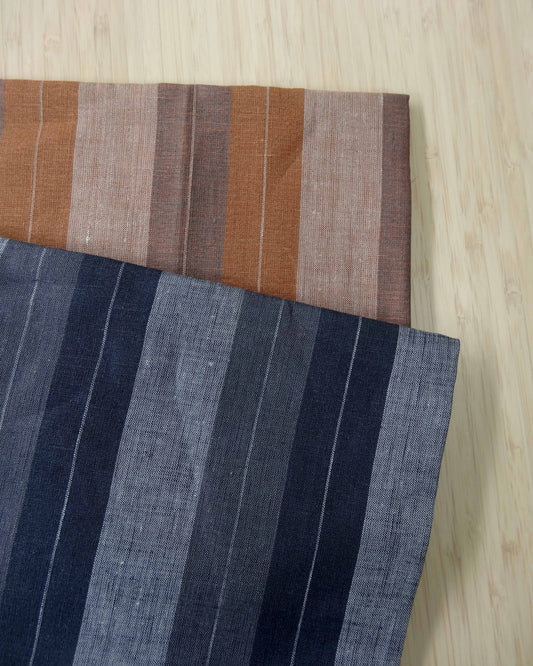 Photo of 2 different colours of striped linen fabrics folded and stacked. One is in orange tones and the other in blue tones.  photographed from above on a wooden table.
