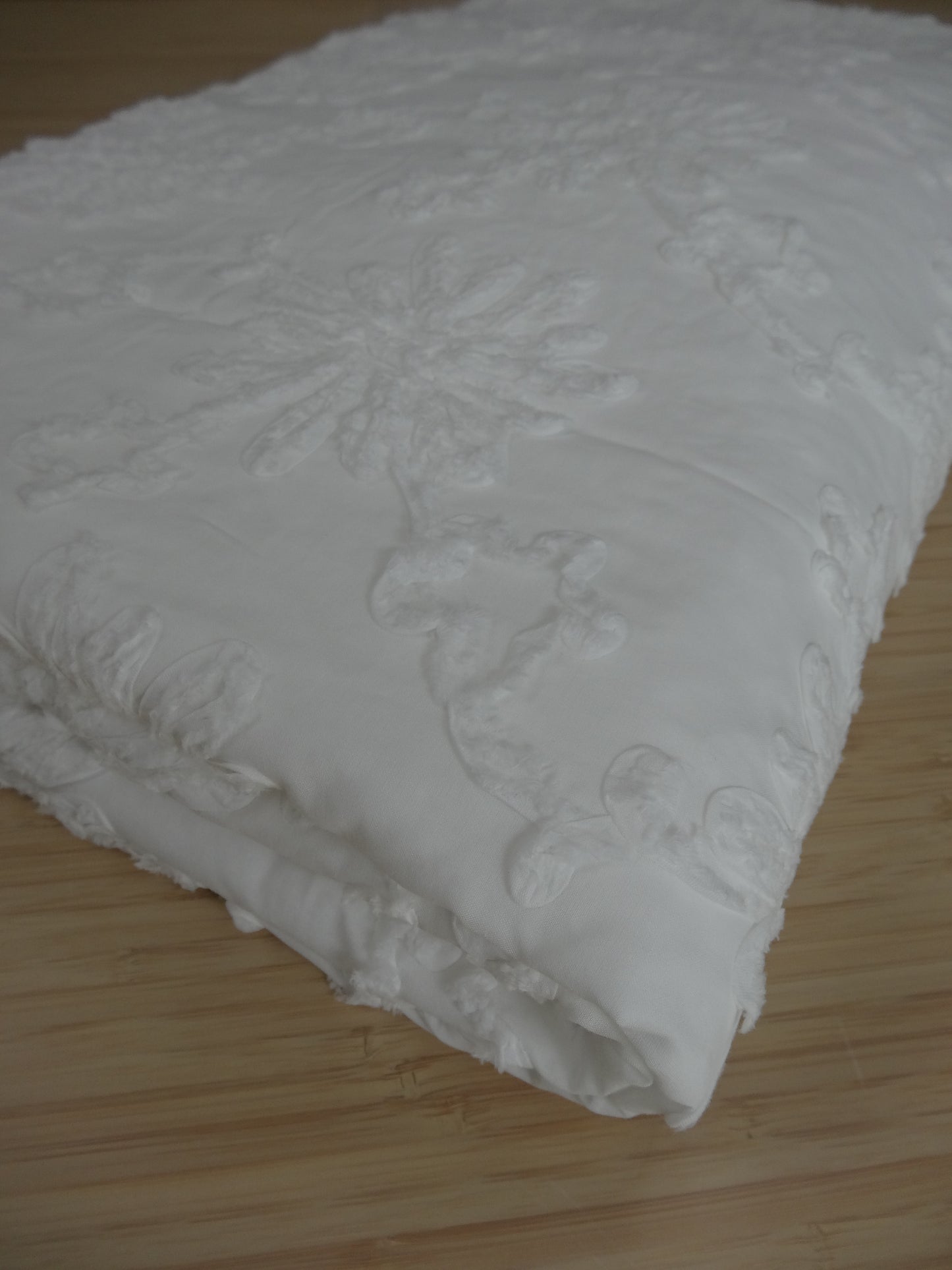 White 100% cotton fabric with white floral applique. Fabric is folded and photographed on a wooden table.