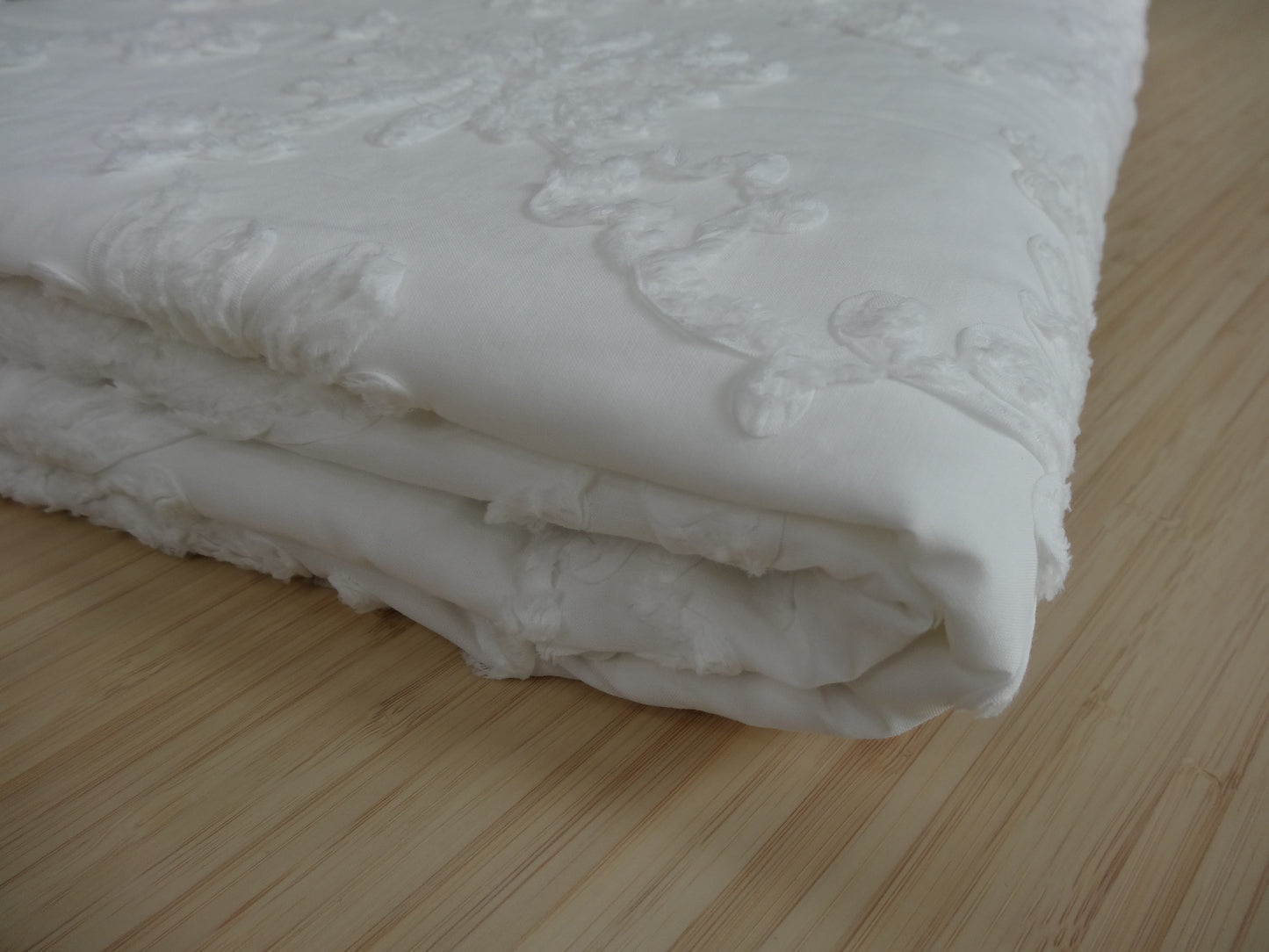 White 100% cotton fabric with white floral applique. It is folded and laid on a wooden table