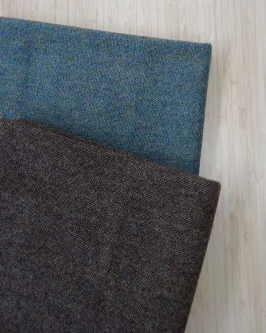 Two pieces of wool fabric folded and stacked. One is a mottled brow and the other is a mottled blue/teal tone. Photographed from above on a wooden table. 
