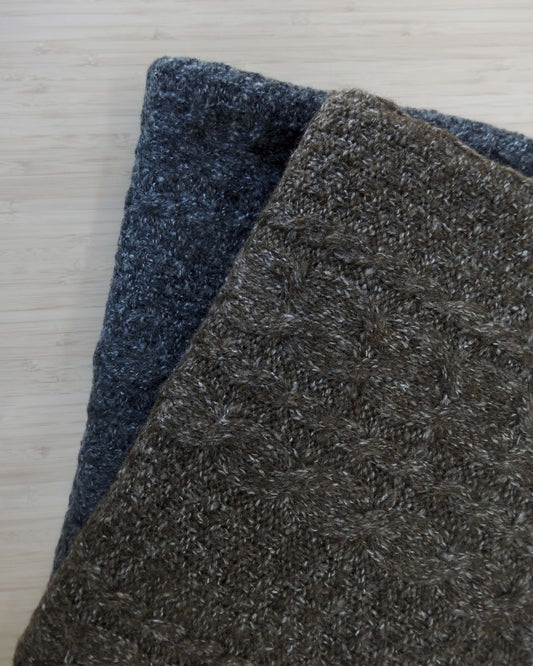 Two folded pieces of wool blend cable knit fabric. A brown and a charcoal grey stacked ontop of each other shot from above on a wooden table