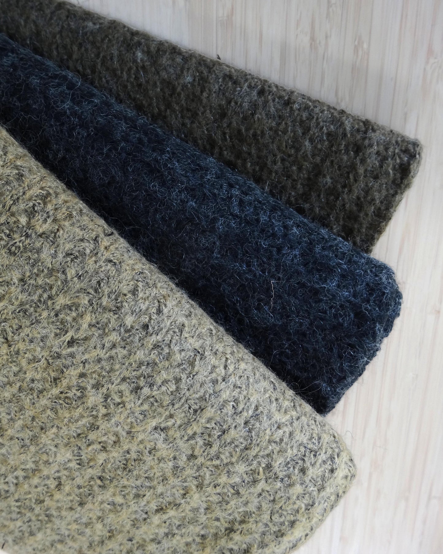 Photo of 3 wool blend fabrics folded and stacked in beige, charcoal grey, and brown photographed from above on a wooden table.