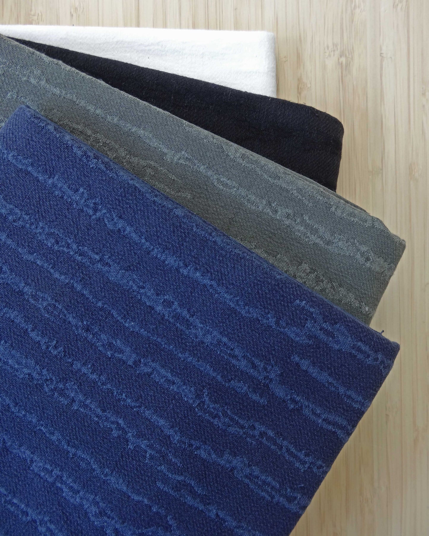 Photo of textured jacquard fabric short ends stacked in blue, khaki green, black, and white on a wooden table. photographed from above.